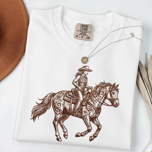 May include: White t-shirt featuring a brown illustration of a person riding a horse, with intricate floral patterns on the horse. A silver necklace with a pendant and a brown hat are also visible.
