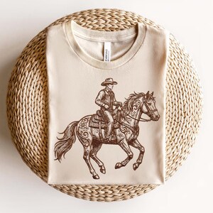 May include: A beige t-shirt featuring a detailed brown illustration of a cowboy riding a horse. The cowboy wears a hat and is seated on a saddle. The horse has decorative floral patterns. The shirt is displayed on a woven, circular surface.