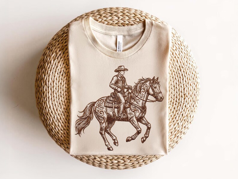 May include: A beige t-shirt featuring a detailed brown illustration of a cowboy riding a horse. The cowboy wears a hat and is seated on a saddle. The horse has decorative floral patterns. The shirt is displayed on a woven, circular surface.
