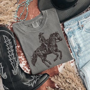 May include: A gray t-shirt featuring a detailed illustration of a cowgirl riding a horse. The image also includes a pair of black cowboy boots, blue jeans, a black hat, and decorative elements.
