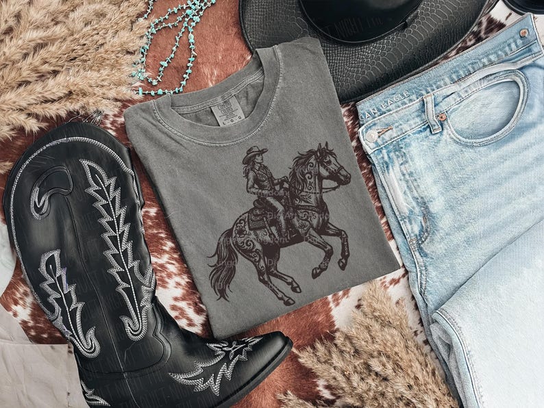May include: A gray t-shirt featuring a detailed illustration of a cowgirl riding a horse. The image also includes a pair of black cowboy boots, blue jeans, a black hat, and decorative elements.