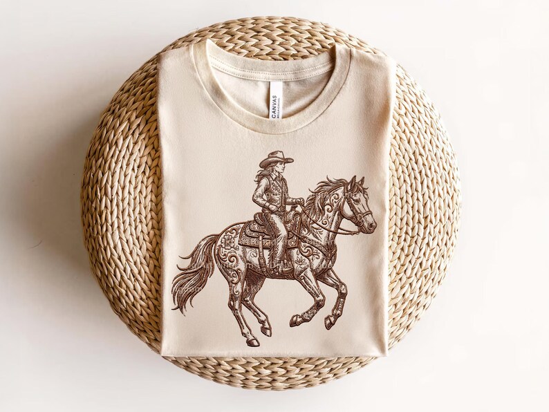 May include: Beige t-shirt featuring a detailed brown illustration of a cowgirl riding a horse. The horse and rider are adorned with intricate patterns. The shirt is displayed on a woven, circular surface.