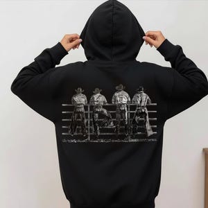 以下が含まれることがあります： 黒色のパーカー。グレーのグラフィックで、柵に寄りかかっている4人のカウボーイが描かれています。カウボーイは帽子とウエスタン風の服装をしています。パーカーには、ドローストリングフードと長袖が付いています。