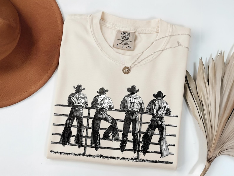 以下が含まれることがあります： クリーム色のTシャツ。白黒で描かれた4人のカウボーイが柵に寄りかかっているイラストが特徴。クルーネックと半袖。小さなペンダントが付いたシルバーのネックレスがTシャツの上に置かれています。