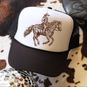 May include: A trucker hat with a brown and white graphic of a person riding a horse. The hat has a white front panel with the graphic, a black mesh back, and a brown brim. The hat is on a cowhide patterned surface.