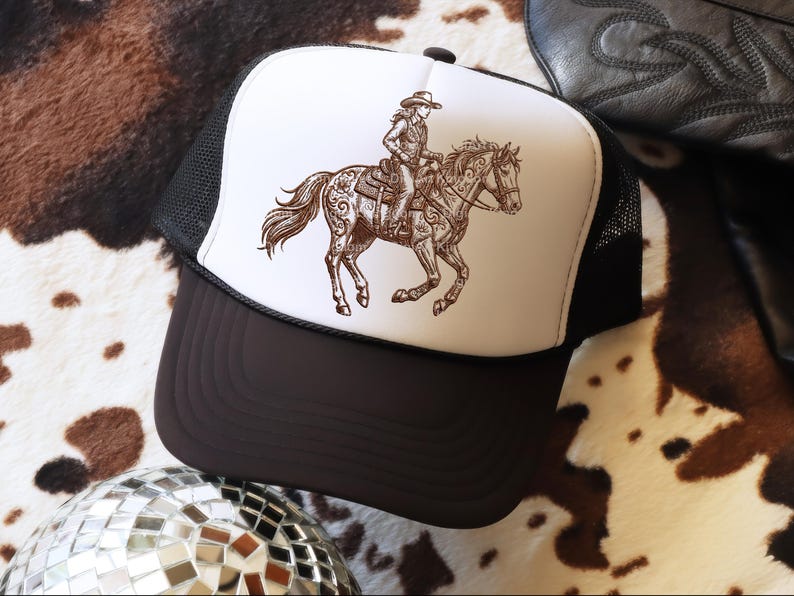 May include: A trucker hat with a brown and white graphic of a person riding a horse. The hat has a white front panel with the graphic, a black mesh back, and a brown brim. The hat is on a cowhide patterned surface.