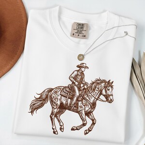 May include: White Comfort Colors t-shirt featuring a brown illustration of a cowboy riding a horse. The cowboy is wearing a hat and riding a horse with ornate saddle details. A silver necklace with a pendant is on top of the shirt.