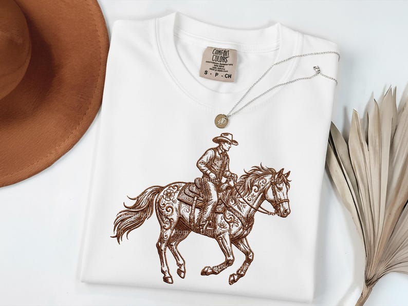 May include: White Comfort Colors t-shirt featuring a brown illustration of a cowboy riding a horse. The cowboy is wearing a hat and riding a horse with ornate saddle details. A silver necklace with a pendant is on top of the shirt.