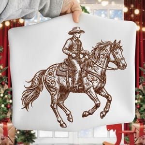 May include: White pillow with a brown illustration of a cowboy riding a horse. The cowboy wears a hat and vest. The horse has decorative floral patterns. The image is held by a person.
