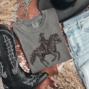 May include: A gray t-shirt featuring a cowboy riding a horse, paired with blue jeans, a black cowboy hat, and black cowboy boots. The boots have white stitching. A turquoise necklace and dried grass complete the western theme.