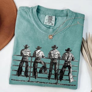以下が含まれることがあります： ターコイズブルーのComfort Colors Tシャツ。4人のカウボーイが柵に寄りかかっているグラフィックが描かれています。カウボーイは帽子とジーンズを着用。ペンダント付きのシルバーネックレスがシャツの上にあります。茶色の帽子と乾燥した植物が背景にあります。
