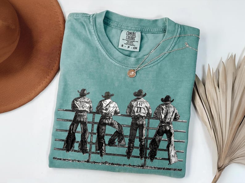 以下が含まれることがあります： ターコイズブルーのComfort Colors Tシャツ。4人のカウボーイが柵に寄りかかっているグラフィックが描かれています。カウボーイは帽子とジーンズを着用。ペンダント付きのシルバーネックレスがシャツの上にあります。茶色の帽子と乾燥した植物が背景にあります。