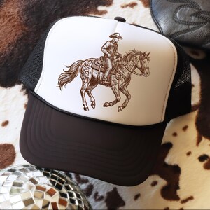 May include: A trucker hat with a brown and white graphic of a cowboy riding a horse. The hat has a white front panel, a black mesh back, and a brown brim. The cowboy and horse are detailed with intricate designs.