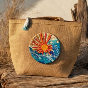 Op de afbeelding: Een beige jute tas met een ronde punch needle geborduurde patch op de voorzijde. De patch toont een heldere gele en oranje zon met stralen boven gestileerde blauwe en turquoise oceaangolven. Er hangt een kleine wit-blauwe kwast aan de rits.