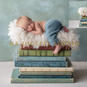 Puede incluir: Un recién nacido dormido, con pantalones azules, descansa sobre una superficie blanca y esponjosa, encima de una pila de libros antiguos. Los libros son de varios colores, incluyendo verde, azul y marrón. El fondo es azul verdoso.