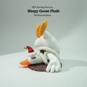 Op de afbeelding: Een pluche gans speelgoed met een kerstmuts en een witte sjaal. De gans heeft een oranje snavel en gele accenten, liggend op een patroonbasis. De tekst "Sleepy Goose Plush" en "ThePatternHaus" zijn zichtbaar.