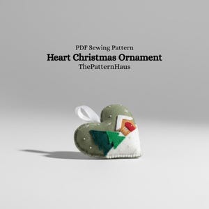 May include: A heart-shaped Christmas ornament with a white ribbon for hanging. The ornament is olive green with white accents, featuring a house, trees, and snowflakes. Text on the image reads "Heart Christmas Ornament" and "ThePatternHaus".