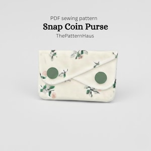 Pode incluir: Uma carteira de moedas com fecho de pressão creme com um padrão floral e dois fechos de pressão verdes. O texto "Snap Coin Purse" é exibido acima da carteira, com "ThePatternHaus" abaixo.