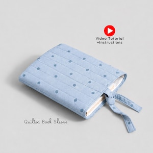Puede incluir: Una funda de libro acolchada de color azul claro con un patrón de pequeños puntos azul oscuro, costuras horizontales y un cierre de lazo. El texto "Quilted Book Sleeve" y "Video Tutorial +Instructions" con un icono de play rojo.