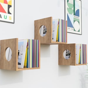May include: Three square wooden wall-mounted shelves with a light brown wood grain finish. Each shelf features a circular cutout on the side panel and holds a collection of colorful vinyl records or thin books arranged vertically inside the frame.