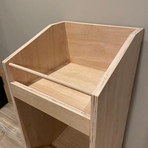 May include: A light-colored, unfinished wooden display shelf with a slanted top and multiple shelves. The shelf has a simple, minimalist design, with a horizontal bar on the front of the top shelf and a similar bar on the front of the lower shelf.