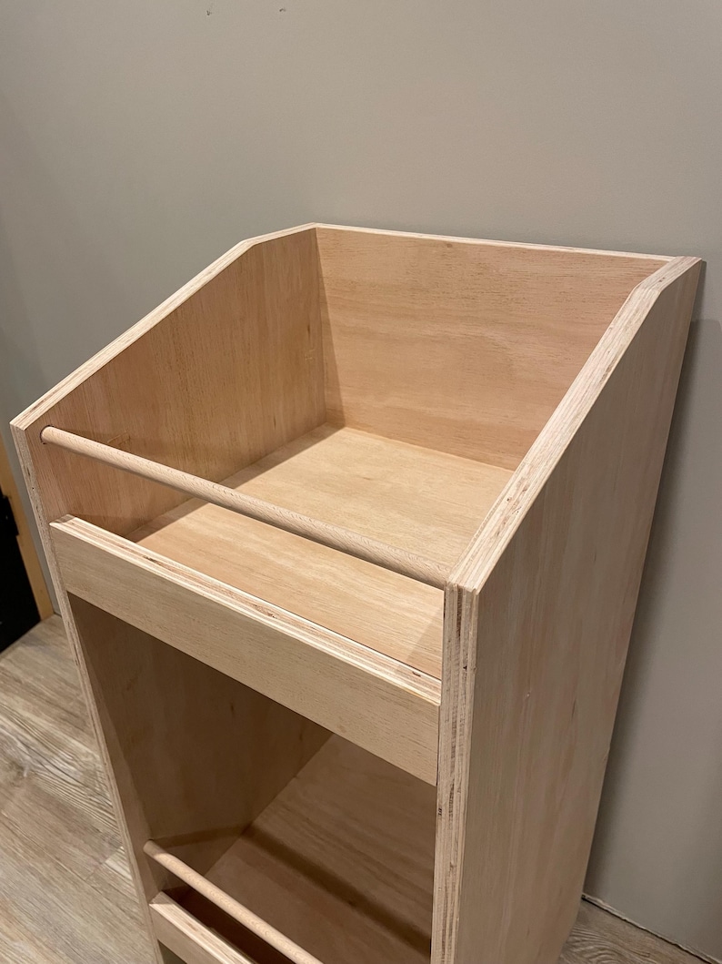 May include: A light-colored, unfinished wooden display shelf with a slanted top and multiple shelves. The shelf has a simple, minimalist design, with a horizontal bar on the front of the top shelf and a similar bar on the front of the lower shelf.