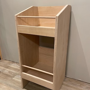 May include: A light-colored wooden bookshelf with two shelves and a sloped top. The bookshelf has two wooden dowels that act as stoppers. The unit is made of plywood and is designed to hold books or other items.