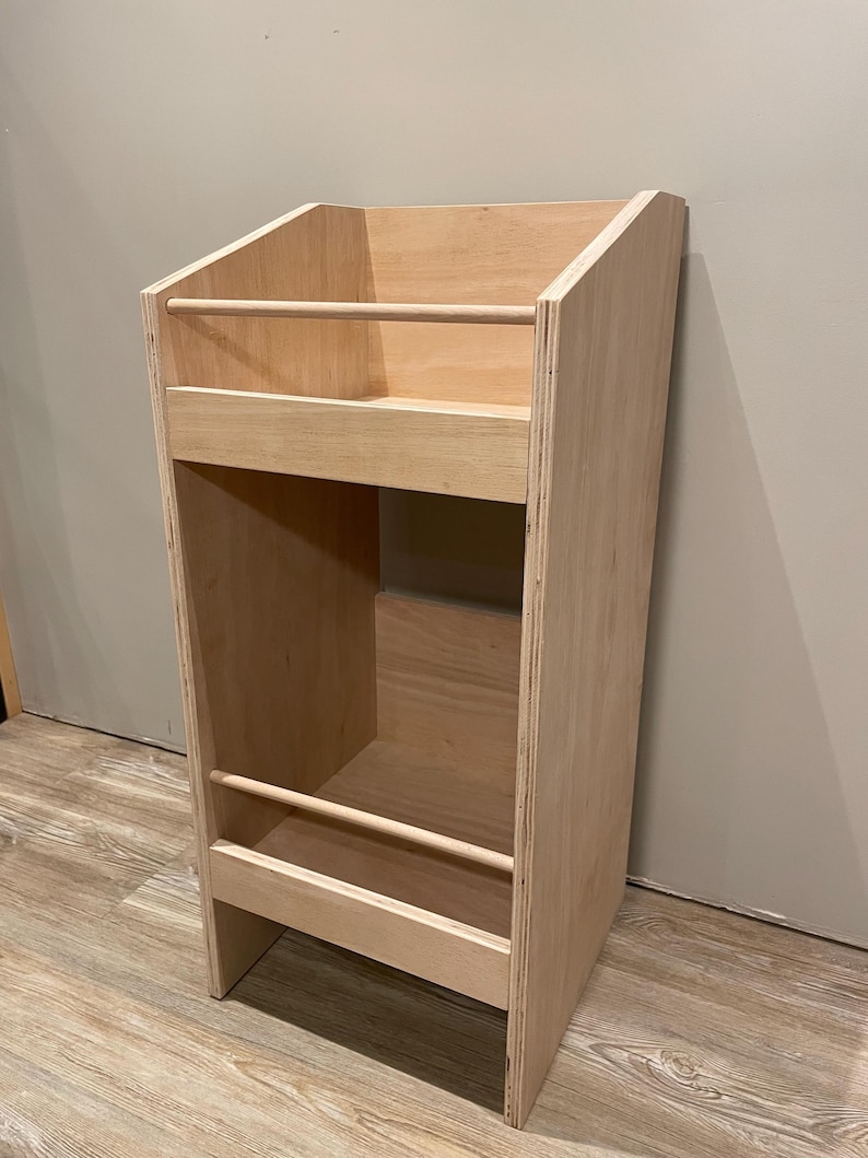 May include: A light-colored wooden bookshelf with two shelves and a sloped top. The bookshelf has two wooden dowels that act as stoppers. The unit is made of plywood and is designed to hold books or other items.
