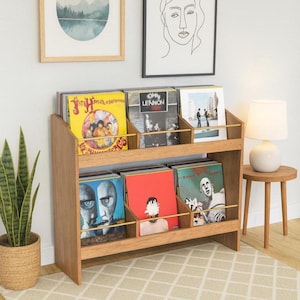 K&ouml;nnte beinhalten: Helles Holz-Schallplattenregal mit zwei Regalb&ouml;den, gef&uuml;llt mit Vinyl-Schallplatten. Das Regal hat eine helle Holzoberfl&auml;che und goldfarbene Trennw&auml;nde. Die Alben haben farbenfrohe Cover, darunter Gelb, Blau und Rot.