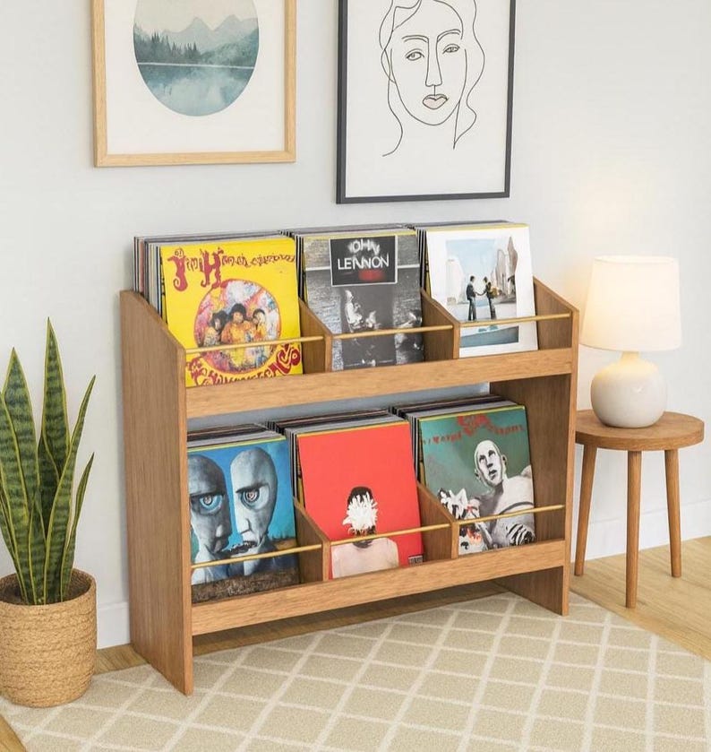 K&ouml;nnte beinhalten: Helles Holz-Schallplattenregal mit zwei Regalb&ouml;den, gef&uuml;llt mit Vinyl-Schallplatten. Das Regal hat eine helle Holzoberfl&auml;che und goldfarbene Trennw&auml;nde. Die Alben haben farbenfrohe Cover, darunter Gelb, Blau und Rot.