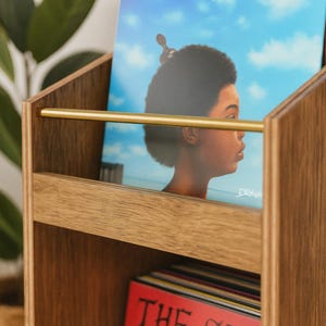 May include: Wooden record holder with a brass bar. The holder contains vinyl records, including one with a portrait against a blue sky and another with the text "THE CLASH" on a red background. The holder is made of light brown wood.
