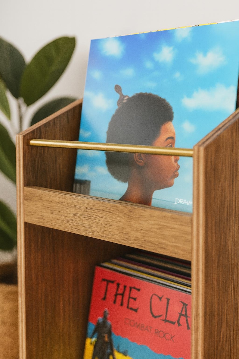 May include: Wooden record holder with a brass bar. The holder contains vinyl records, including one with a portrait against a blue sky and another with the text "THE CLASH" on a red background. The holder is made of light brown wood.