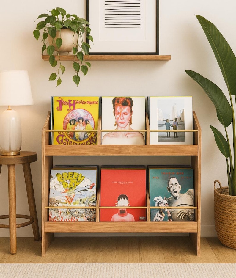 K&ouml;nnte beinhalten: Ein h&ouml;lzernes Schallplattenregal mit sechs Vinyl-Alben. Die Alben haben farbenfrohe Cover, darunter ein gelbes Album mit einem Bandfoto, ein Portr&auml;t von David Bowie und ein Green Day-Album. Eine Lampe und Pflanzen sind ebenfalls im Bild.