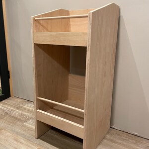 May include: A light-colored wooden bookshelf with a rectangular design. It features two shelves with wooden dowels and a tall, open storage area. The bookshelf is made of plywood and has a natural wood finish.