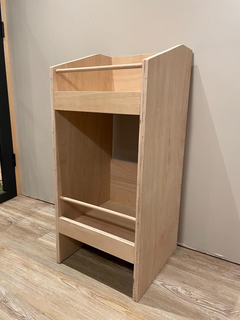 May include: A light-colored wooden bookshelf with a rectangular design. It features two shelves with wooden dowels and a tall, open storage area. The bookshelf is made of plywood and has a natural wood finish.