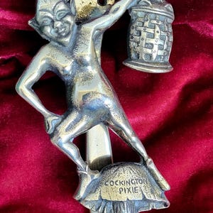 May include: A silver door knocker depicting a pixie holding a lantern. The pixie stands on a mushroom with the words "Cockington Pixie" engraved. The knocker is designed for mounting and is made of metal.