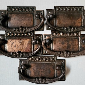May include: Five antique bronze drawer pulls. Each has a rectangular backplate and a curved handle. The pulls have a decorative design and a dark, aged patina, suggesting vintage or repurposed use.