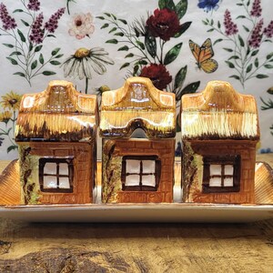 May include: A set of three ceramic house-shaped containers arranged on a rectangular tray. Each house features a brown brick-textured facade, a white window with a brown frame, and a glossy tan roof. The tray has a matching brown border.