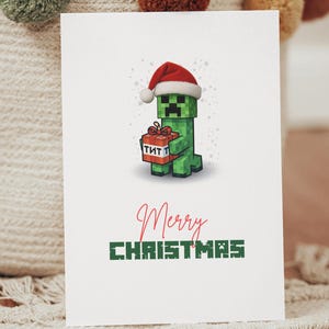 Puede incluir: Una tarjeta de Navidad blanca con un personaje verde pixelado con un gorro de Papá Noel y un regalo de TNT. La tarjeta tiene las palabras "Merry Christmas" en escritura roja y letras pixeladas verdes.