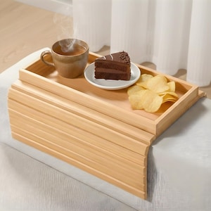 May include: A light wooden lap tray holding a cup of tea, a slice of chocolate cake on a white plate, and potato crisps. The tray has a curved design. The tray is resting on a light grey surface.