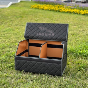 May include: Black quilted car organizer with a diamond pattern. The interior features tan compartments and black straps. The organizer is open, revealing the interior, and sits on a grassy surface.