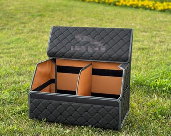 May include: Black quilted car organizer with a diamond pattern. The interior features tan compartments and black straps. The organizer is open, revealing the interior, and sits on a grassy surface.