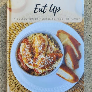 May include: A cookbook titled "Eat Up" with the subtitle "A Collection of Recipes to Feed the Family." The cover shows a bowl of pasta with chicken and toasted bread. The authors are Katie Muonio and Rebecca Swisse.