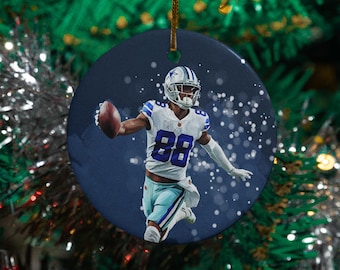 May include: A round Christmas ornament featuring a football player in a white and blue uniform, holding a football. The ornament has a dark blue background with white accents and a gold string for hanging.