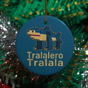May include: A round, blue ceramic Christmas ornament with a pixelated shark design in blue, tan, and white. The text "Tralalero Tralala" is printed in gold below the shark. A gold string is attached for hanging.