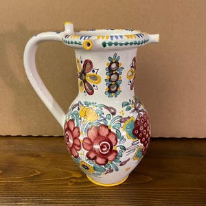 May include: A white ceramic pitcher featuring a hand-painted floral pattern in red, yellow, and green. The vessel has a curved handle, a wide rim with small decorative protrusions, and a rounded body decorated with clusters of grapes and flowers.
