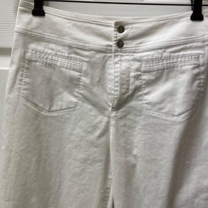 May include: A pair of white pants featuring a high-rise waistband with two visible buttons at the center front. The garment includes two large patch pockets on the front, reinforced with horizontal stitching along the top edges and a central fly closure.
