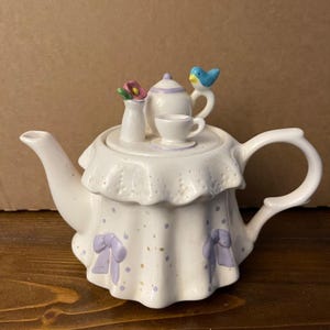Op de afbeelding: Een witte keramische theepot met een geplooide rand, paarse stippen en lichtpaarse strikken. Het deksel is versierd met een miniatuur theepot, een kopje op een schoteltje, een vaasje met een roze bloem en een blauw vogeltje op het handvat.