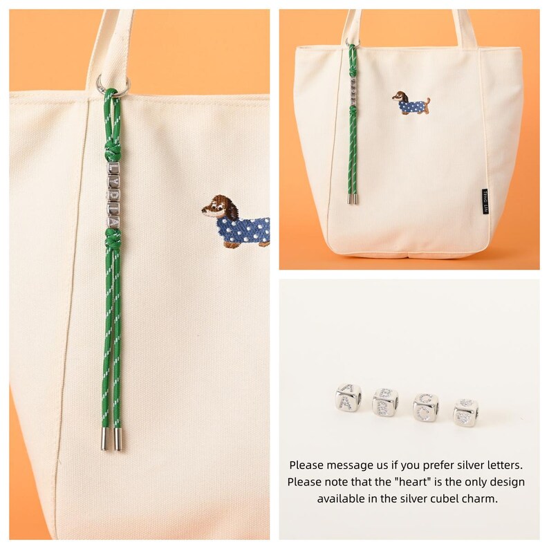 May include: Cream-colored tote bag with a green and white rope handle and a dachshund dog embroidered on the front. The dog is wearing a blue coat with white polka dots. A green rope with silver letter charms hangs from the handle.