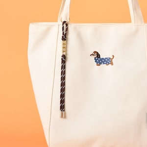 May include: Cream-colored tote bag with a brown and white rope detail and a gold-colored charm. The bag features an embroidered dachshund wearing a blue polka-dot sweater. The bag has two handles.